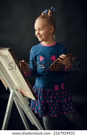A happy smiling young girl with blond hair, in a blue dress, stands near an easel and paints with a brush in a dark creative studio.