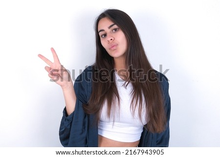young caucasian woman wearing fashion clothes over white background makes peace gesture keeps lips folded shows v sign. Body language concept