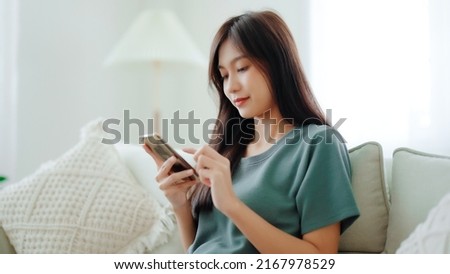 Happy young asian woman relax on comfortable couch at home texting messaging on smartphone, smiling girl use cellphone chatting, browse wireless internet on gadget, shopping online from home Royalty-Free Stock Photo #2167978529