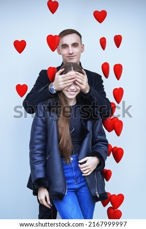Valentine's day. Handsome young man closes the eyes of a joyful, smiling girl. Happy young people in love. White background with hearts decorations. Love and relationship. 