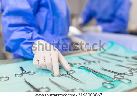 Nurse hand taking surgical instrument for group of surgeons at background operating patient in surgical theatre. Steel medical instruments ready to be used. Surgery and emergency concept Royalty-Free Stock Photo #2168008867