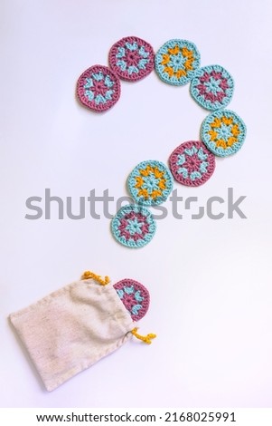 Question mark made of circle crochet motifs with a dot coming out of a cloth bag on white background. Crochet patterns of turosque, lilac and yellow.