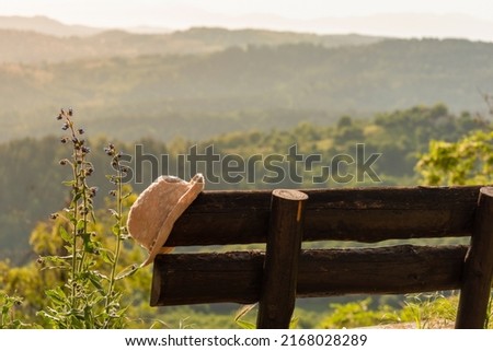 Hat on wooden rustic bench against background of nature mountains and atmospheric epic forest valley. Peaceful, quiet, serene summer evening in sunset light while travel, vacation or weekend getaway Royalty-Free Stock Photo #2168028289