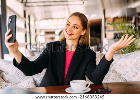 Photo of a happy smiling brunette young woman indoors in cafe drinking coffee using mobile phone take a selfie.