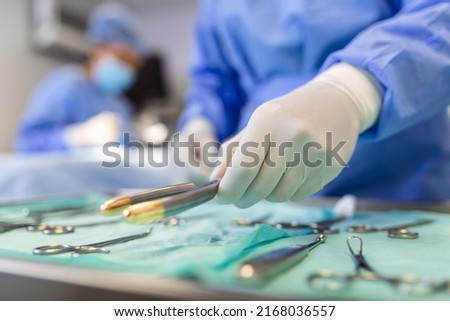 Nurse hand taking surgical instrument for group of surgeons at background operating patient in surgical theatre. Steel medical instruments ready to be used. Surgery and emergency concept Royalty-Free Stock Photo #2168036557
