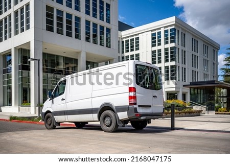Commercial white compact cargo mini van for local delivery and small business standing on the urban city street with multilevel residential apartment buildings serving neighborhood Royalty-Free Stock Photo #2168047175