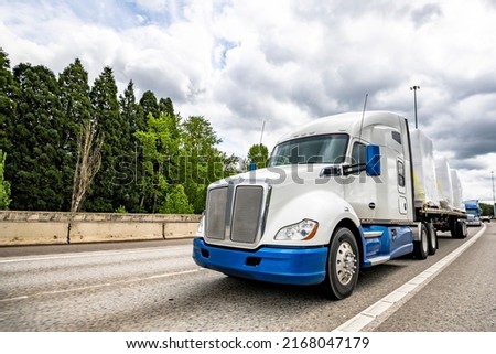 Blue and white big rig bonnet industrial semi truck tractor transporting covered fastened commercial cargo on flat bed semi trailer driving for delivery on the wide multiline turning highway road