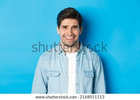 Close-up of young man feeling awkward, smile and cringe from uncomfortable situation, standing over blue background Royalty-Free Stock Photo #2168051113