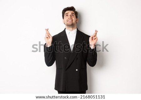 Portrait of tensed and worried handsome businessman, crossing fingers and looking up, begging god, making a wish, standing against white background in black suit