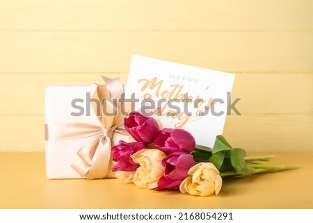 Card with text HAPPY MOTHER'S DAY, gift box and bouquet of tulips on wooden yellow background
