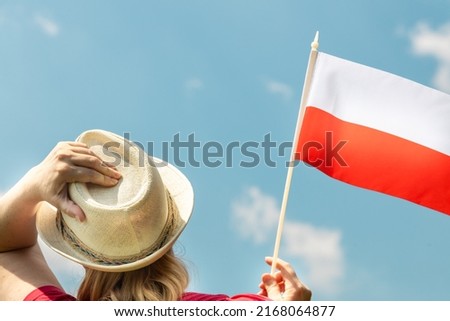 A young woman proudly holds the Polish flag above her head, against the sky with her other hand holding her hat, Concept of national holidays and patriotic events Royalty-Free Stock Photo #2168064877