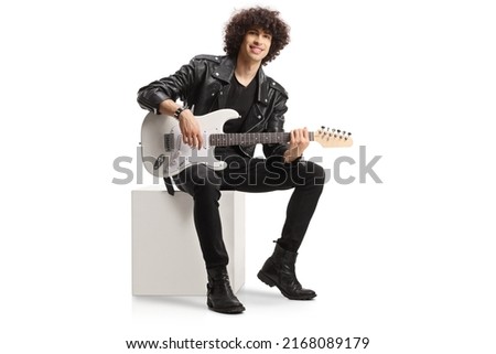 Young male rocker in a leather jacket sitting on a white cube with an electric guitar isolated on white background Royalty-Free Stock Photo #2168089179