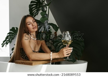 Beautiful fit young woman in a leopard print swimsuit with a glass of white wine in the bathroom. Happy weekend