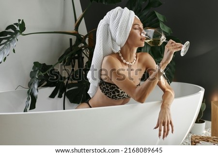 Beautiful young woman in a leopard print swimsuit drinks wine in the bathroom. The girl enjoys a glass of white wine.