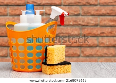 Plastic basket with bottles of dishwashing liquid, glass and tile cleaner, detergent for microwave ovens and stoves, sponges with a brick wall on the background. Washing and cleaning concept.
