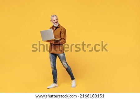 Full size body length fun businesslike elderly gray-haired bearded man 50s years old wears brown shirt go move hold use work on laptop pc computer isolated on plain yellow background studio portrait