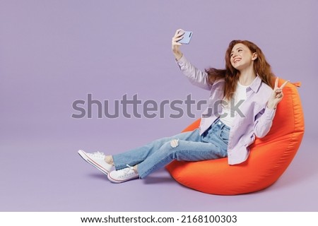 Full size body length young redhead curly woman 20s wears white T-shirt violet jacket sit in bag chair do selfie shot on mobile cell phone isolated pastel purple color wall background studio portrait