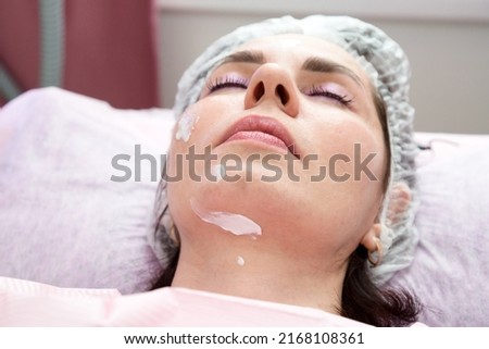 An adult brunette woman with her eyes closed after a facial rejuvenation procedure with cream on her face.