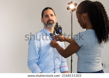 Putting Lavalier Microphone on Caucasian Male Wearing Business Attire. Video Production or Film Set Behind the Scenes. Mic Clips Onto Shirt. Lighting Seen in Background