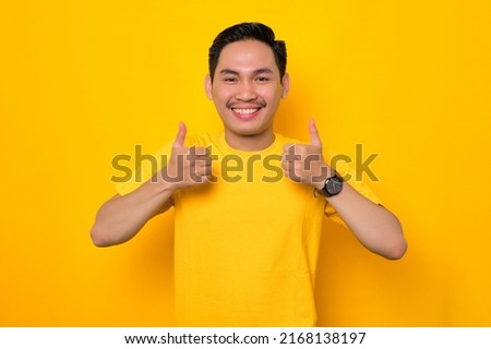 Smiling young Asian man in casual t-shirt showing thumbs up gesture, approving something good isolated on yellow background. People lifestyle concept