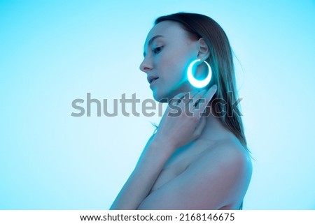 Portrait of tender young girl with neon colored accessories, earrings in shape of circle posing isolated over blue background. Concept of beauty, creativity, art, digitalization, fashion trends