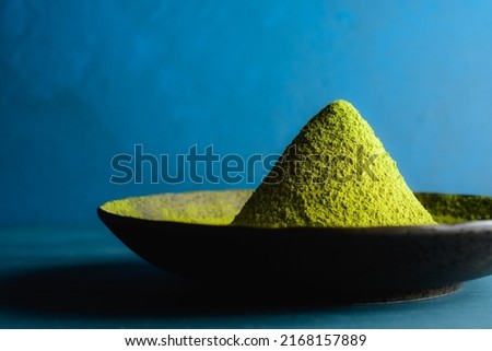 Japanese traditional green tea (matcha) powder in a heap on a black round plate against a blue design board.