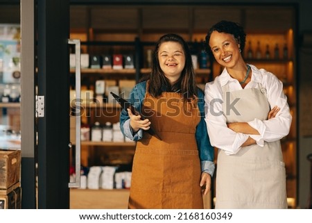 Two successful store employees smiling at the camera while standing at the entrance of their grocery store. Happy woman with Down syndrome working in a local supermarket with her female colleague. Royalty-Free Stock Photo #2168160349