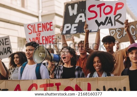 Group of multicultural teenage activists protesting against war and violence in the city streets. Generation Z demonstrators shouting slogans and raising banners while marching for world peace.