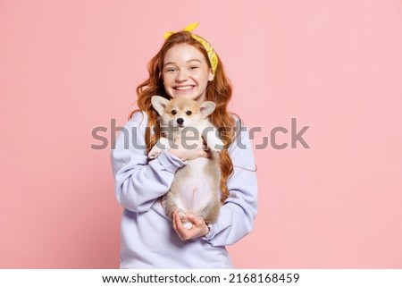Hugs. Cheerful redhead young girl with long curly hair holding cute little puppy of corgi dog isolated on pink background. Care, support and love. Concept of youth, beauty, life. Copy space for ad