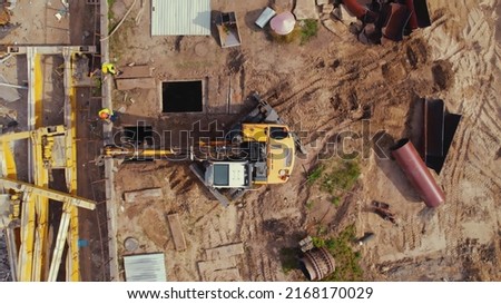 Aerial shot of a construction site. Workers dressed in high-visibility jackets supervising the work of an excavator digging a hole. High quality photo