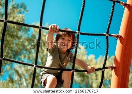 A child climbs up an alpine grid in a park on a playground on a hot summer day. children's playground in a public park, entertainment and recreation for children, mountaineering training. Royalty-Free Stock Photo #2168170717