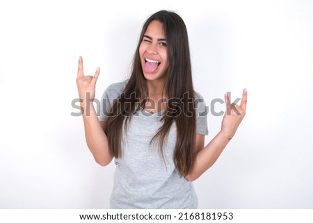 young caucasian woman wearing grey t-shirt over white background making rock hand gesture and showing tongue
