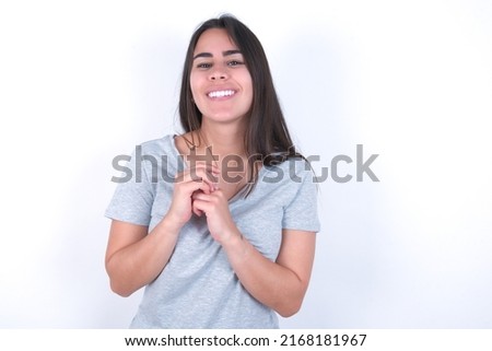 Positive young caucasian woman wearing grey t-shirt over white background smiles happily, glad to receive pleasant news from interlocutor, keeps hands together. People emotions concept.