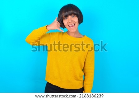 Young woman with bob haircut wearing yellow sweater over blue wall makes phone gesture, says call me back again, has glad expression.