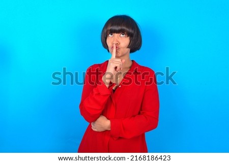 Young woman with bob haircut wearing red shirt over blue wall  makes silence gesture, keeps index finger to lips makes hush sign. Asks not to share secret.