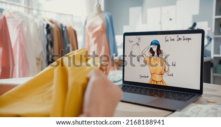 Close-up young Asia lady dressmaker with laptop on graphic sketch photo use tape measure fabric compare sample create dress design new collection on desk in shop. Tailor startup business concept.