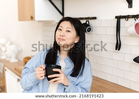 An attractive Asian woman rests in the kitchen, holds a mug with a hot drink and looks into the camera.
