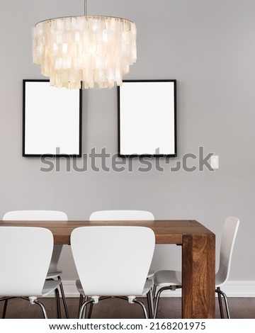 A modern dining room with a chandelier hanging above a wooden table and white chairs with empty picture frames on the wall.