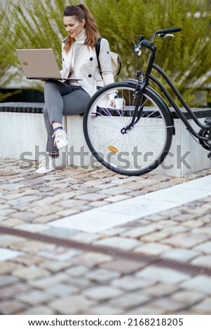modern woman in beige trench coat with bicycle and laptop sitting on the bench outdoors on the city street.