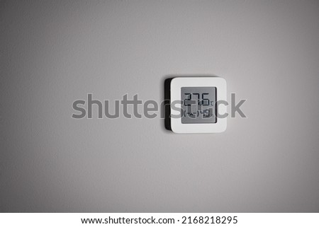 Electronic indoor white plastic digital thermometer on gray wall showing temperature, humidity level in house, smiling smiley face indoors in daylight sun