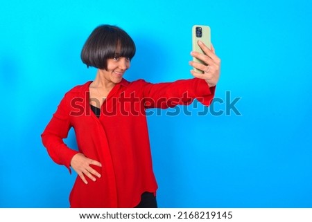 Isolated shot of pleased cheerful Young woman with bob haircut wearing red shirt over blue wall, makes selfie with mobile phone. People, technology and leisure concept
