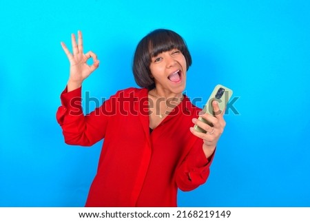 Happy Young woman with bob haircut wearing red shirt over blue wall sending a message on his smartphone or taking a selfie  and making ok sign with his hand.