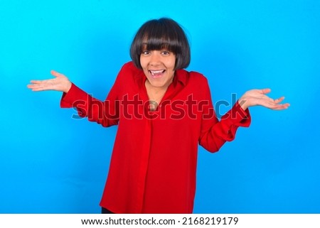So what? Portrait of arrogant Young woman with bob haircut wearing red shirt over blue wall shrugging hands sideways smiling gasping indifferent, telling something obvious. Royalty-Free Stock Photo #2168219179