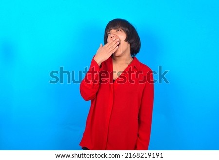 Sleepy Young woman with bob haircut wearing red shirt over blue wall yawning with messy hair, feeling tired after sleepless night, yawning, covering mouth with palm.