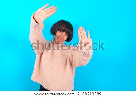 Portrait of smiling Young woman with bob haircut wearing pink knitted sweater shirt over blue wall looking at camera and gesturing finger frame. Creativity and photography concept.