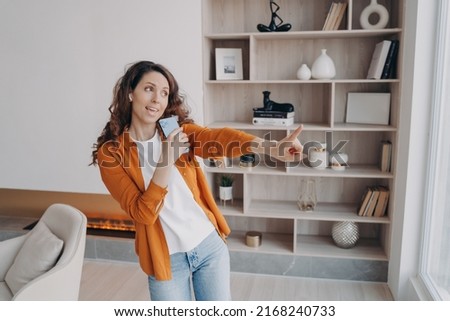 Weekend morning at home. Emotional caucasian woman is singing with phone as with microphone. Carefree girl having fun in her living room in modern apartment. Electronic gadgets using.