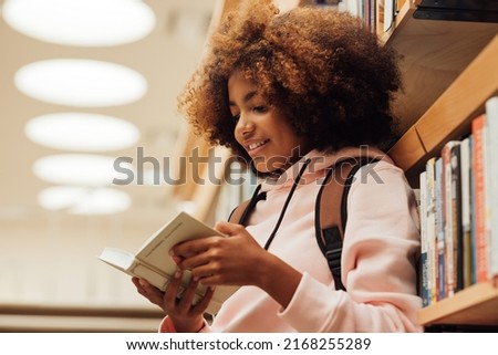 Girl in casuals with backpack leaning bookshelf in library. Student reading a book while standing in library.