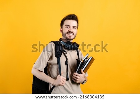 Portrait of man wearing beige tee posing isolated over yellow background student with books and backpack and looking at the camera. High school university college concept.
