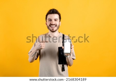 Happy casual man wearing t-shirt posing isolated over yellow background showing to camera and pointing at device. Gadget with empty free space for mock up. White blank screen.