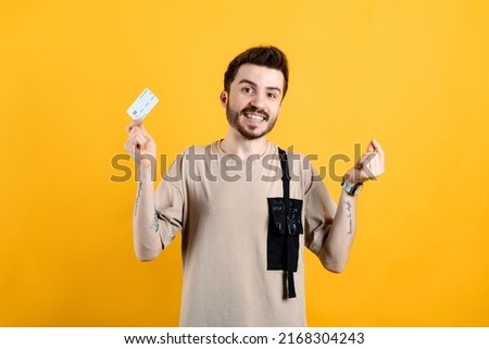 Cheerful young man wearing t-shirt posing isolated over yellow background holding credit card and money sign, smiling face and happy earnings